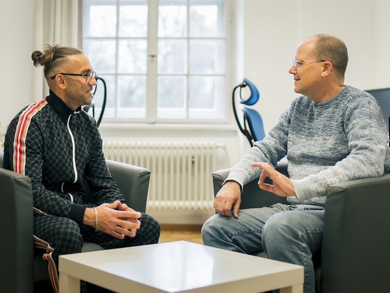 Zwei Männer sitzen in schwarzen Sesseln und unterhalten sich.